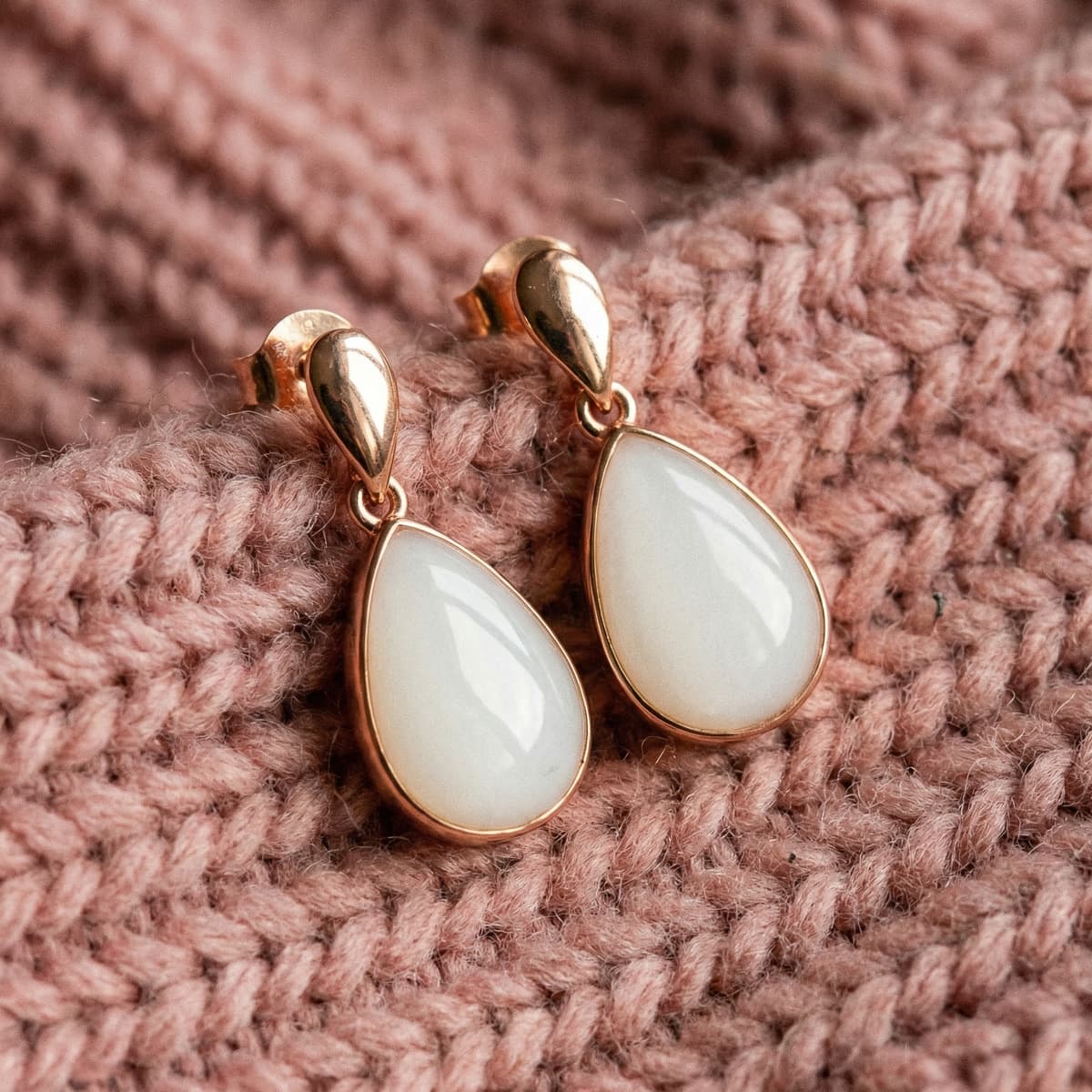 Rose gold teardrop breastmilk keepsake earrings with creamy white resin stones on a soft pink knitted background, showcasing results from a breastmilk earrings diy making kit.