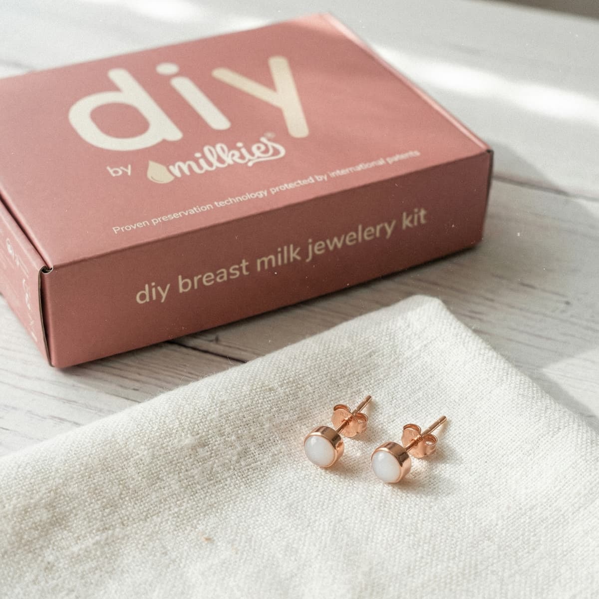 Rose-gold stud earrings on a linen cloth next to a DIY by MILKIES box showing a breastmilk earrings making kit for creating breast milk jewelry at home.