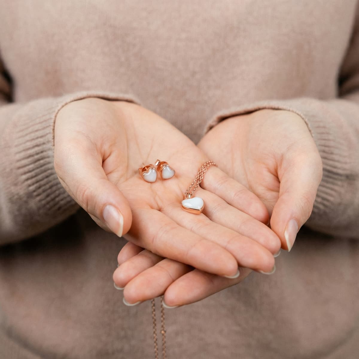 Hands holding a rose-gold heart pendant necklace and matching heart stud earrings, showcasing keepsakes made using a breastmilk jewellery making kit.