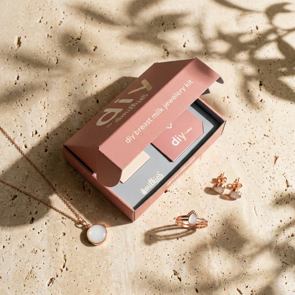 Pink DIY by MILKIES box on a stone surface displaying a breastmilk jewellery making kit with rose-gold necklace, ring, and earrings featuring milky white resin stones.