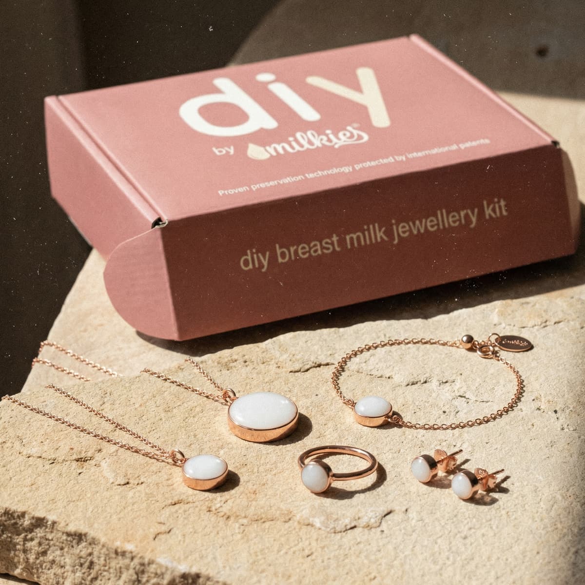 Rose-gold plated DIY by MILKIES breastmilk jewelry diy making kit box with necklace pendants, bracelet, ring, and stud earrings featuring white breastmilk resin stones on a beige stone surface.