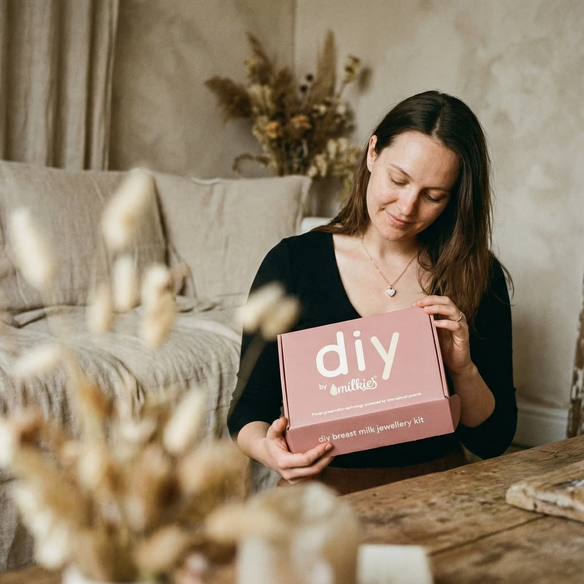 Mother at home holding a DIY by MILKIES box with a breastmilk jewelry making kit, highlighting an easy, private way to create a personal keepsake using patented preservation technology.