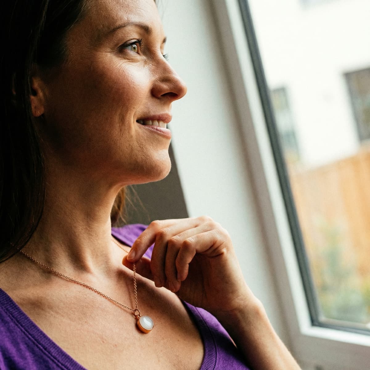 Smiling mom standing by a window wearing an oval keepsake pendant on a delicate rose-gold chain, showing the finished result from a breastmilk necklace making kit.