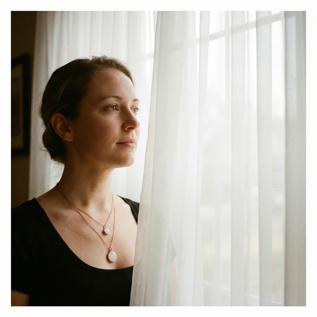 Woman wearing layered breastmilk jewelry by a window, showcasing finished pendants from a breastmilk necklaces making kit in gold-toned settings with milky white resin stones.