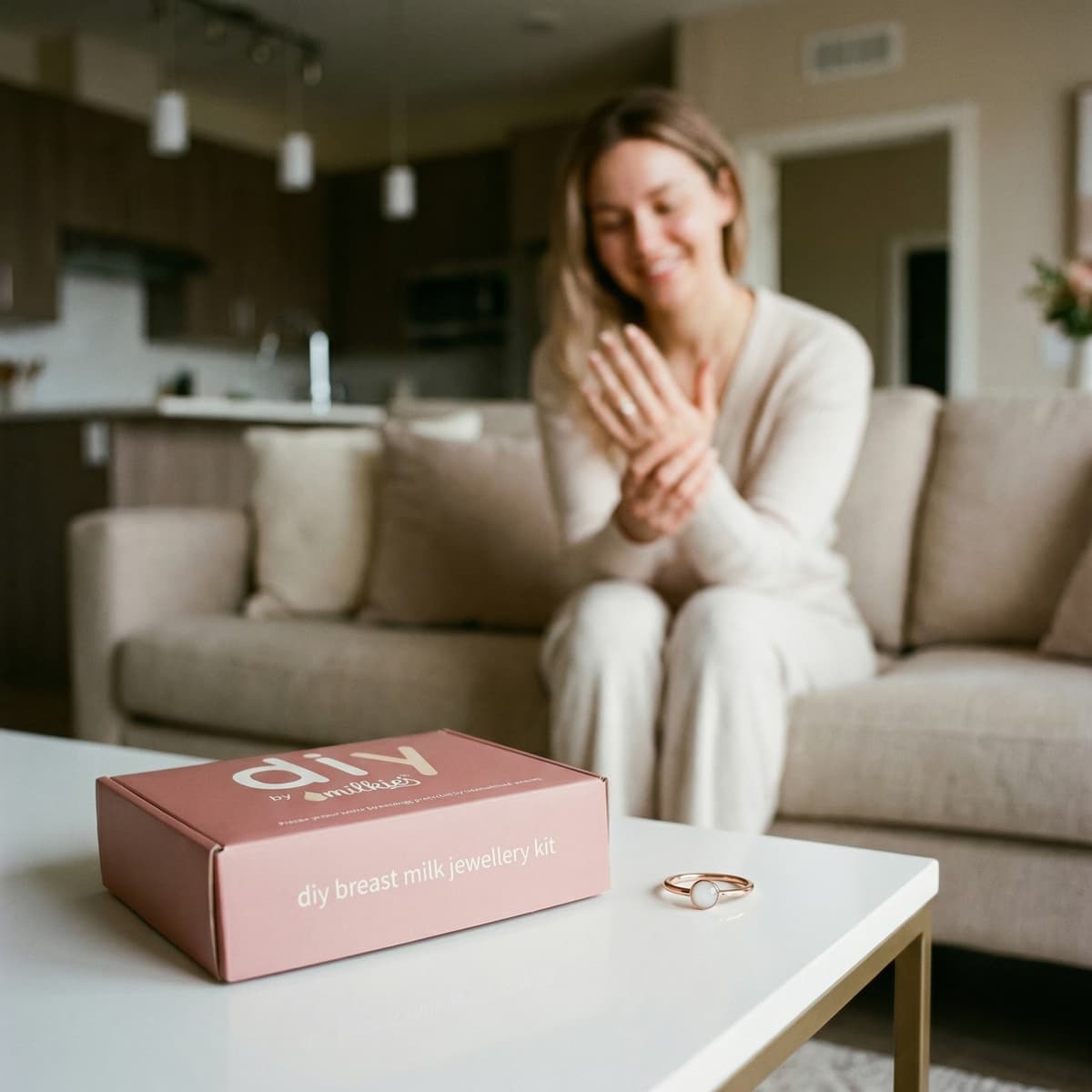 Pink MILKIES box labeled “diy breast milk jewellery kit” on a coffee table beside a ring, with a smiling woman on a sofa admiring her hand in a cozy living room.