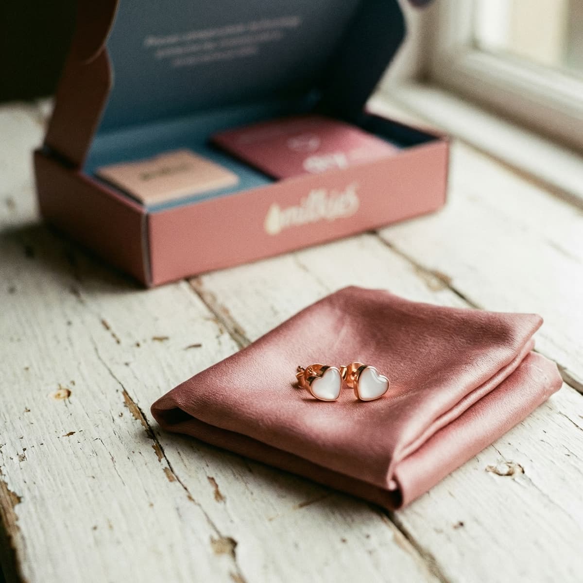 Rose-gold heart stud breastmilk earrings on blush fabric with an open DIY by MILKIES box in the background, highlighting an at-home diy breastmilk earring making kit for creating a keepsake.