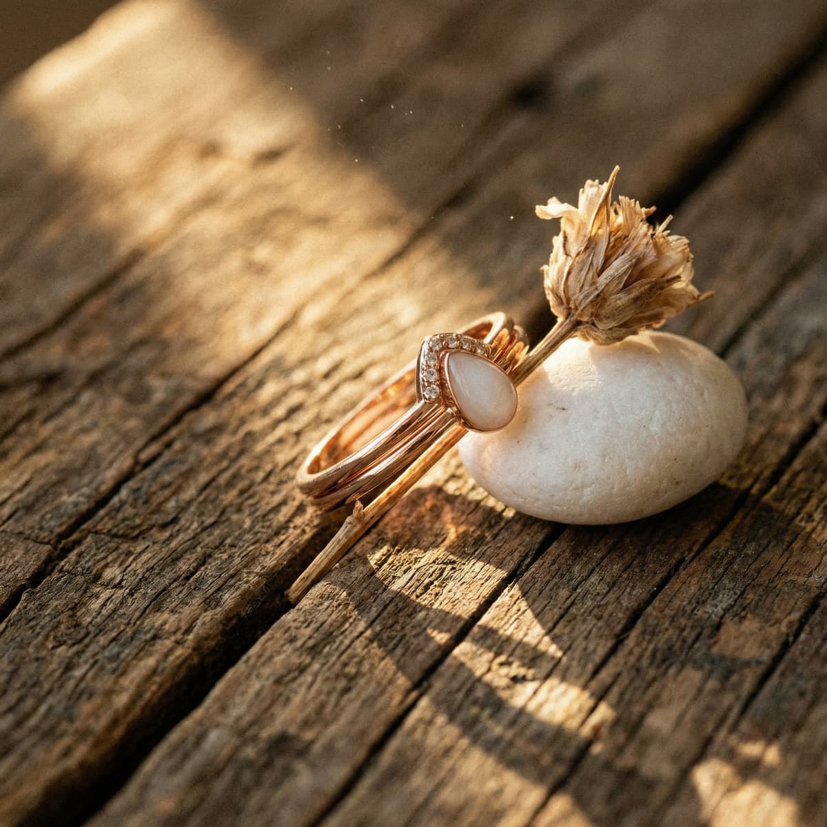 Rose gold ring with a milky white teardrop stone on rustic wooden boards beside a dried flower and white pebble, showcasing a diy breastmilk ring making kit keepsake.