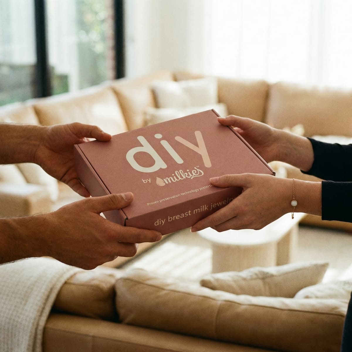 Hands exchanging a DIY by MILKIES breast milk jewelry kit box in a cozy living room, a thoughtful gift for new mother’s day gift that lets moms create a personalized keepsake at home.