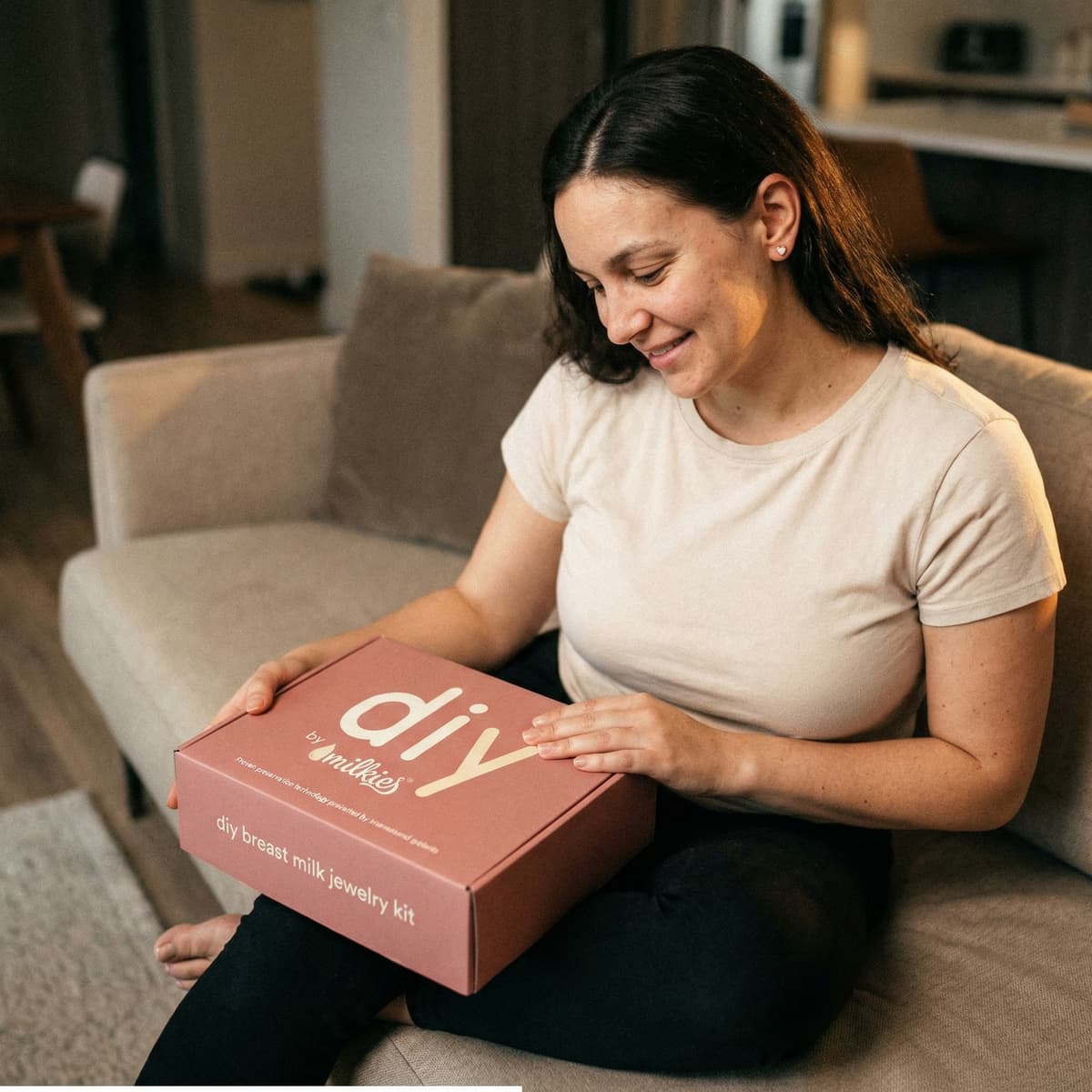 Mom smiling on a sofa while opening the DIY by MILKIES breast milk jewelry keepsake kit at home, a thoughtful gift for new mother’s day gift that shows why an easy at-home kit is a great idea.